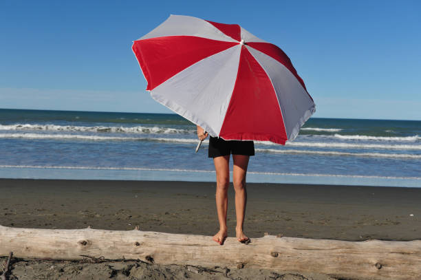 Young fit woman stands covering her face under colorful parasol on a sandy beach.