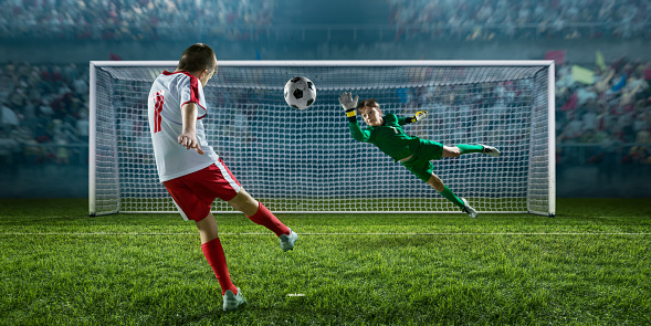 Soccer kids player scoring a goal. Goalkeeper tries to hit the ball. In the foreground there is a football gate. Players are dressed in a professional football uniform. Behind them is a stadium with fans in the stands.