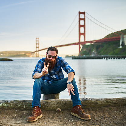 bearded man making peace sign and looking at camera in front of the golden gate bridge