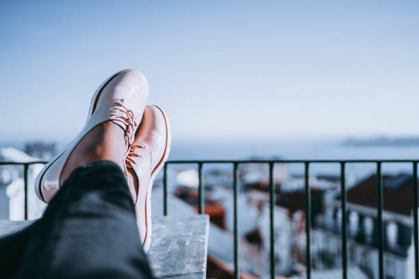 Feet of resting girl with glossy pale pink shoes on the wooden table and touristic resort city with coast and multiple houses in defocused background with copy space for advertising, logo or your text