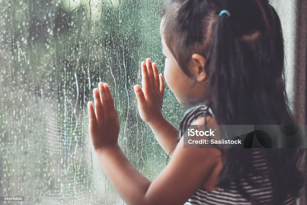 Triste niña asiática mirando afuera por la ventana en el día de lluvia - Foto de stock de Lluvia libre de derechos