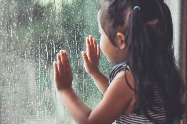triste bambina asiatica che guarda fuori dalla finestra nella giornata piovosa - stagione delle piogge foto e immagini stock