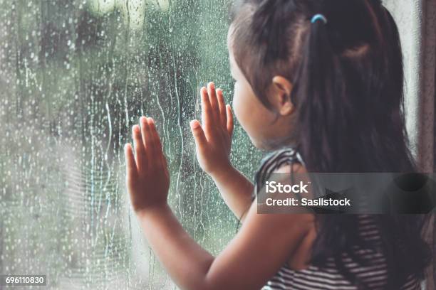 Sad Asian Little Girl Looking Outside Through The Window In The Rainy Day Stock Photo - Download Image Now