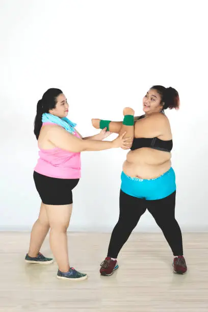 Portrait of fat woman stretching her friend hands while exercising together in the gym center