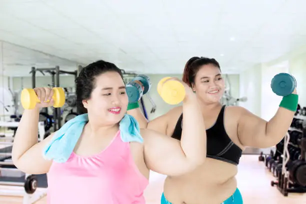 Two women lifting dumbbells while training together and standing in the gym center