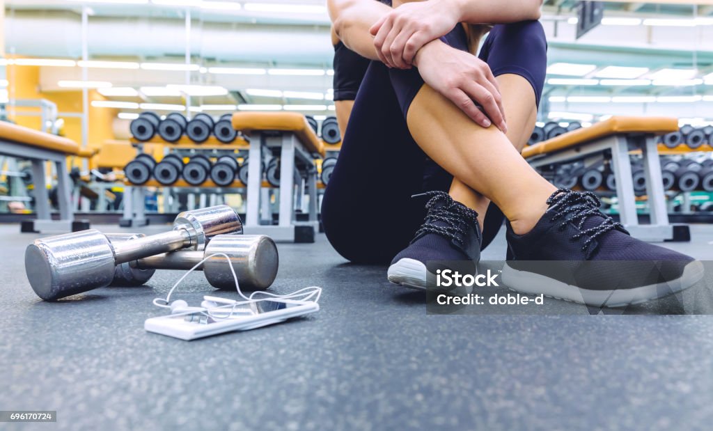 Deportivo mujer sentada con mancuernas y teléfono inteligente en el gimnasio del piso - Foto de stock de Centro de bienestar libre de derechos