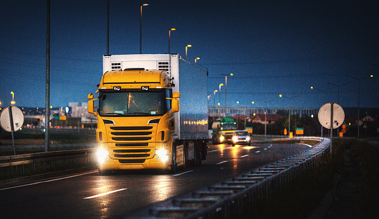 Closeup front view of a yellow cargo truck driving under on a highway at late sunset. It's leaving the city . Some unrecognizable cars are driving behind.
