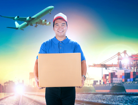 delivery man holding card box toothy smiling face against shipping port and cargo plane flying background