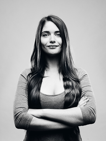 Black and white portrait of confident beautiful young woman standing with happy expression. Real people female is against gray background with big smile and arms crossed. She has long brown hair. Vertical studio photography from a DSLR camera. Sharp focus on eyes.