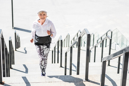 Portrait of happy old woman raising upstairs. She is jogging on city ladder construction and smiling