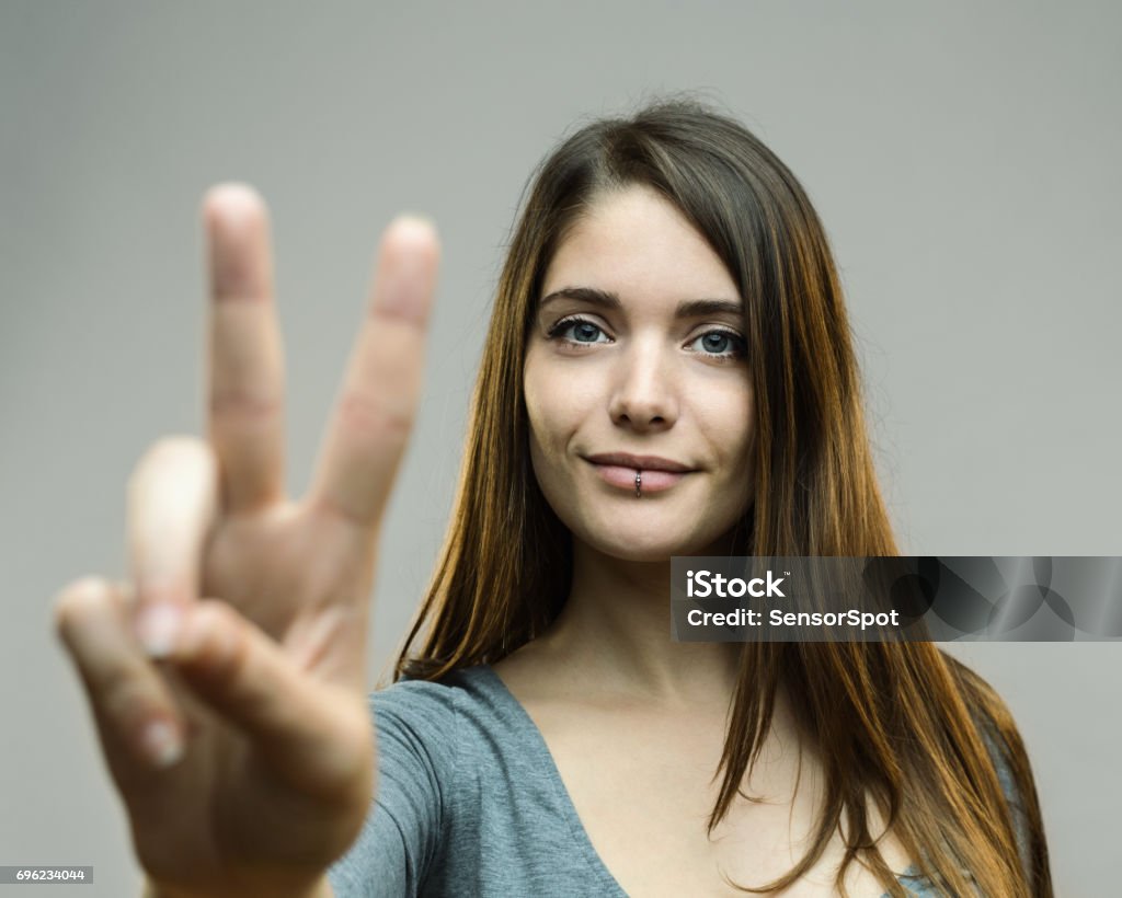 Real woman showing victory sign Portrait of young woman gesturing victory sign against gray background. Horizontal shot of real woman showing peace hand sign. Studio photography from a DSLR camera. Sharp focus on eyes. Peace Sign - Gesture Stock Photo