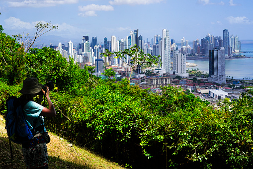 Senior woman loking at Panama City with its skyscrapers from Ancon hill, the highest hill in Panama City. To the view point there is 30 min. hiking through remains of with city isolated Jungle. In front is green jungle and on top of photo is blue sky with white clouds. Panama City is the financial and shipping hub of Central America.
