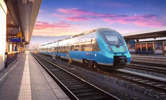 Beautiful view with modern commuter train on the railway station and colorful sky with clouds at sunset in Europe. Industrial landscape with blue train on railway platform. Railroad background