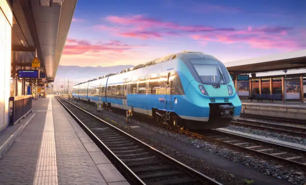 Photo of Beautiful view with modern commuter train on the railway station and colorful sky with clouds at sunset in Europe. Industrial landscape with blue train on railway platform. Railroad background
