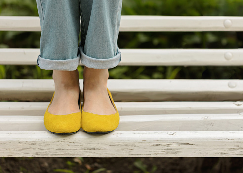 Female legs in blue jeans and yellow shoes on a white bench.