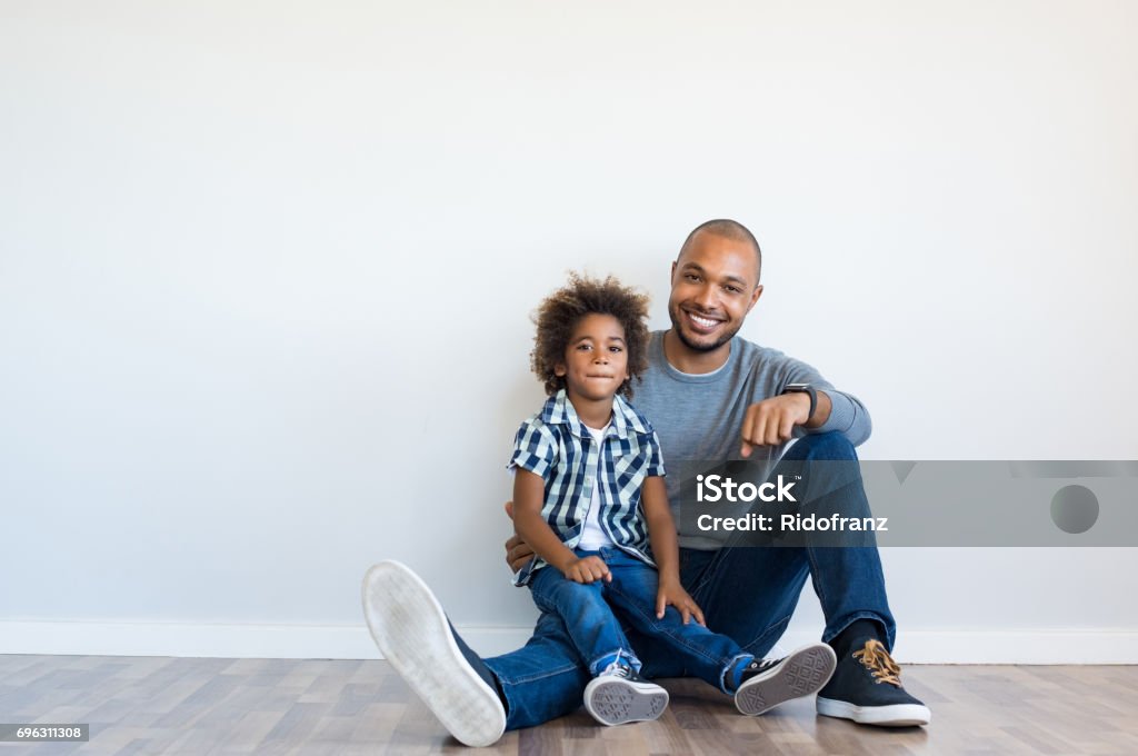 Happy father and son sitting Cheerful black father sitting on floor with smiling son and leaning on blank wall with copy space. Young african boy sitting on father lap in a new home. Happy father and child sitting and looking at camera. Father Stock Photo