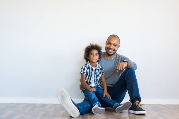 Happy father and son sitting Cheerful black father sitting on floor with smiling son and leaning on blank wall with copy space. Young african boy sitting on father lap in a new home. Happy father and child sitting and looking at camera. african father stock pictures, royalty-free photos & images