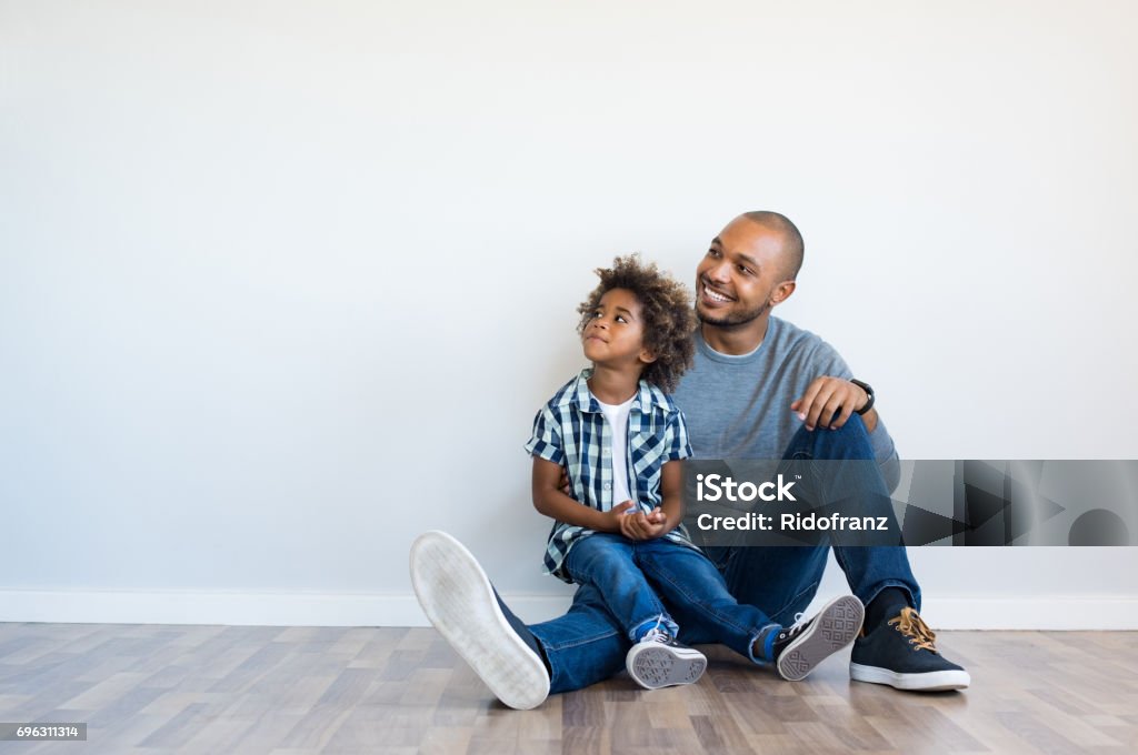 Father and son thinking African father and his son sitting on floor and looking up in a blank wall. Happy dad and little boy sitting in an empty room. Young black man with his child thinking and pensive with copy space. Father Stock Photo