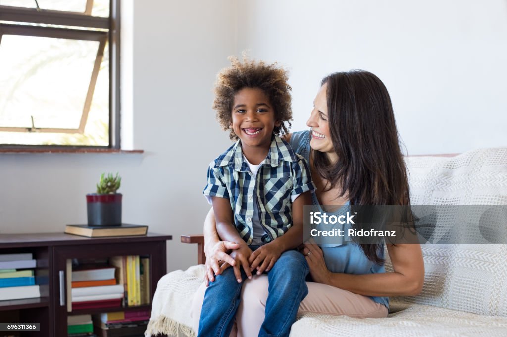 Adopted child playing with mother Happy latin mother embracing her adopted son on couch and smiling. Cheerful woman playing with her african child at home. Portrait of son and mom having fun together after school. Child Care Stock Photo