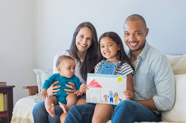 Happy family and new home Daughter showing drawing of a happy family outside a new house. Cute infant looking at colorful drawing of his sister. Happy proud multiethnic parents sitting with children on sofa  and looking at camera. social services photos stock pictures, royalty-free photos & images
