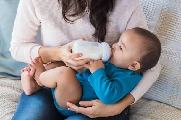 Little infant baby lying on mothers hand drinking milk from bottle. Newborn baby drinking milk from bottle. Cute toddler with milk bottle on leg of mother.