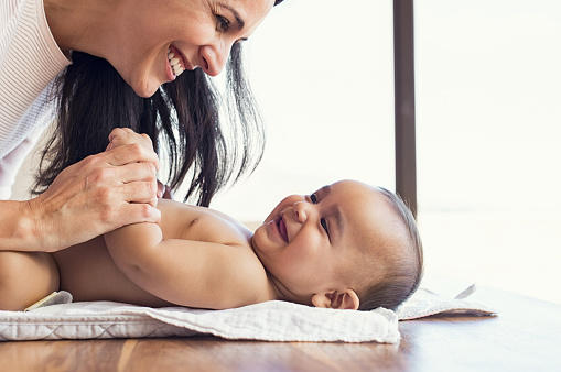 Happy mother playing with baby while changing his diaper. Smiling young woman with baby son on changing table at home. Close up of cheerful mom and toddler boy playing together.