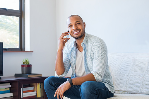 Cheerful black man in a happy conversation over mobile phone. Pensive african guy sitting on sofa talking on cellphone. Smiling man in casual talking on cell phone and thinking while looking up.