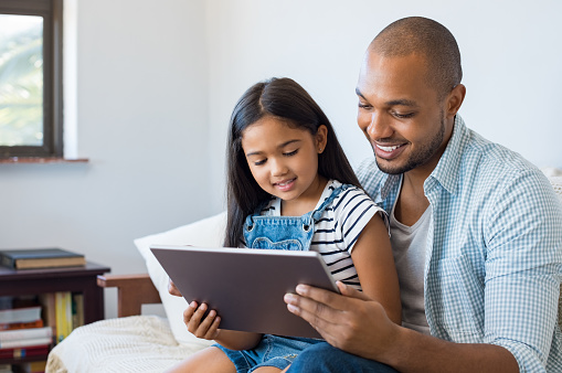 African father and smiling daughter sitting on sofa using digital tablet. Happy dad watching cartoon with his cute little girl. Cheerful father and daughter sitting on couch at home and playing with computer.