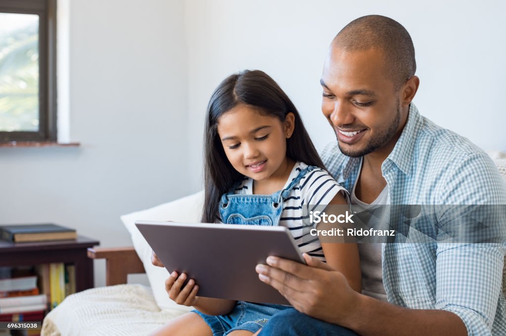 Father and daughter using tablet African father and smiling daughter sitting on sofa using digital tablet. Happy dad watching cartoon with his cute little girl. Cheerful father and daughter sitting on couch at home and playing with computer. Child Stock Photo