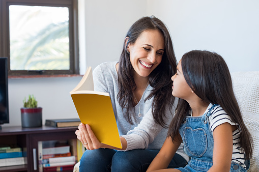 Happy mother reading story book to little girl. Happy latin mother and daughter reading a book while sitting on couch at home. Mother reading a fairy tale to adopted child.