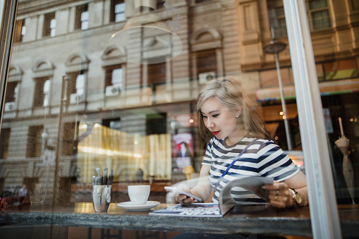 Young Asian woman sitting in a coffee shop reading a magazine and enjoying a cup of coffee.