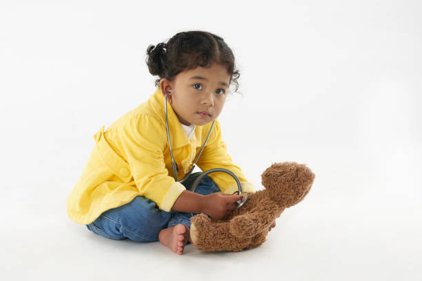 Cute little girl playing doctor and listening to her teddy bear with stethoscope, isolated on white background