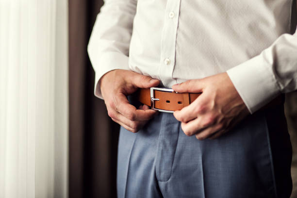 uomo d'affari che si mette una cintura. l'uomo indossa la cintura marrone. concentrati sulla fibbia. sposo che si tiene per mano sulla cintura, abito da sposa - cuff link suit men wedding foto e immagini stock