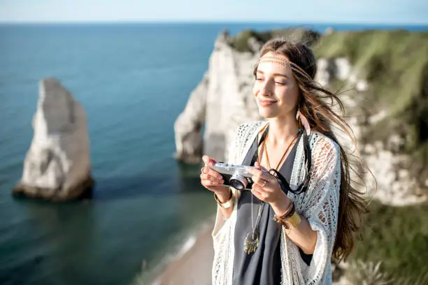 Young woman dressed in hippie style photographing beautiful landscape standing on the rocky coastline in France