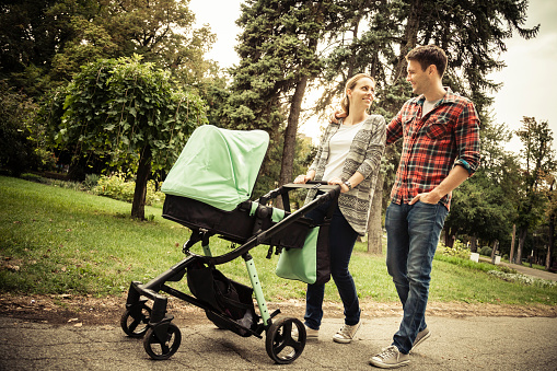 Young urban parents walking in park in the afternoon with their little baby in stroller. The are smiling and happy and enjoy together.