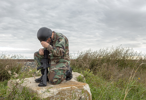 horizontal image of a soldier kneeling down on a big boulder with his gun and his head down saying a prayer.