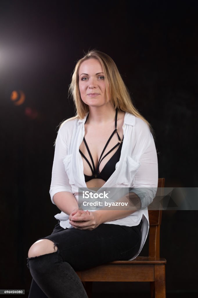 Blond woman on a dark background Beautiful blond woman in her thirties in unbuttoned white shirt that reveals sexy underwear on a dark background at a photo studio. Adult Stock Photo