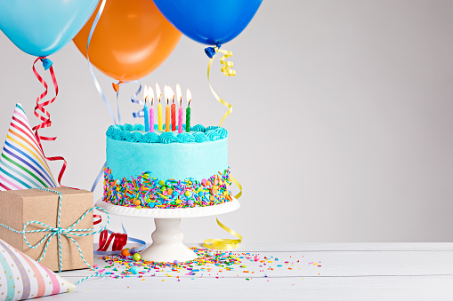 Blue Birthday cake, presents, hats and colorful balloons over light grey.