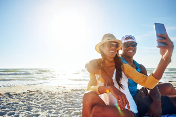 Cheese to memories worth capturing Shot of a happy young couple taking a selfie on the beach cap hat photos stock pictures, royalty-free photos & images