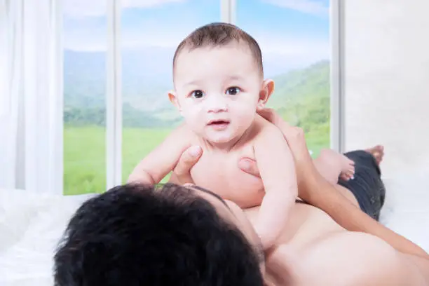 Cute baby boy smiling at camera while lying on father's chest in bedroom with mountain view on the window