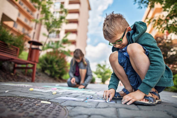 Kids chalking on the pavement in residential area. The boy aged 7 and girl aged 11 are drawing with colorful chalk.\n