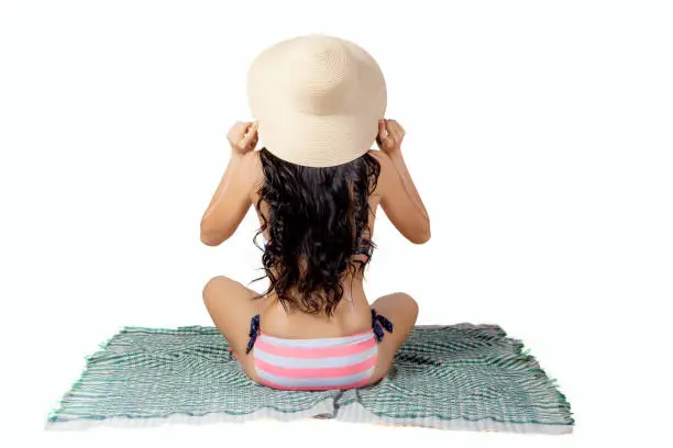 Back view of young woman wearing swimsuit and hat while sitting on the mat, isolated on white background