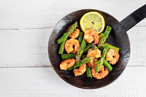 fried prawns or shrimps with  green asparagus peaks and a lemon slice in a black iron pan on rustic white painted wood with copy space, top view from above, selected focus