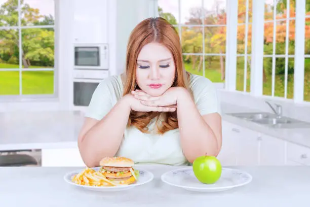 Photo of a blonde woman sitting in the kitchen and looks confused to choose apple fruit or hamburger