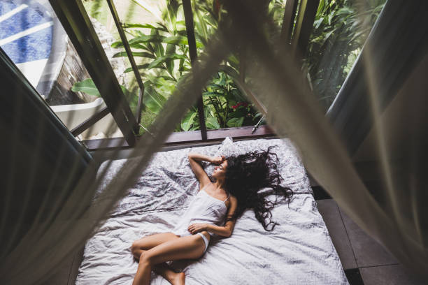 femme en lingerie blanche se prélasser dans son lit le matin, vue depuis la fenêtre sur le jardin tropical. photo de mode de vie - waking up women bedding underwear photos et images de collection