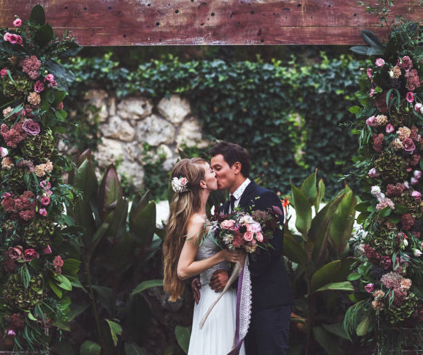 удивительная свадебная церемония с большим количеством свежих цветов в стиле rustic. счастливые молодожены целуются - женатые фотографии стоковые фото и изображения