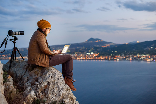 Young attractive photographer sitting on a rock and working on laptop with perfect view of sea, port, night city and sky full of clouds. Camera on tripod beside him.