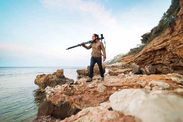 istock Young male photographer walking on rock beach with camera on tripod 696579012