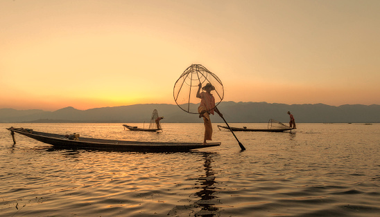 Three fisherman trying to catch fish and posing for camera during in beautiful light of sunset at famous Inle lake of Myanmar in his wooden boat