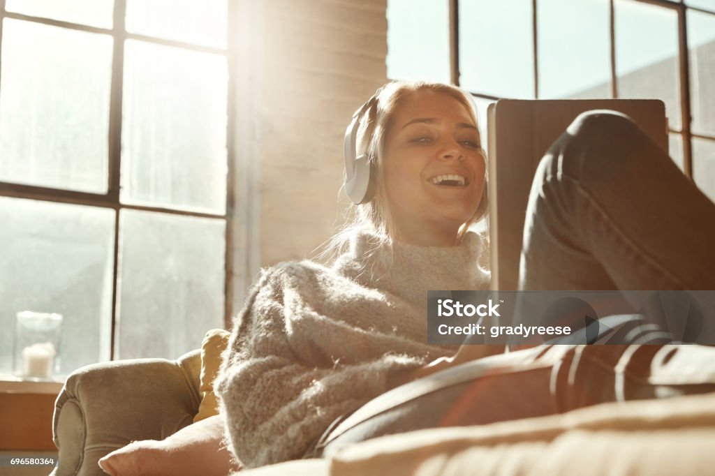 Smart technology's got her entertainment covered Shot of a happy young woman relaxing on the sofa and using headphones with a digital tablet Downloading Stock Photo